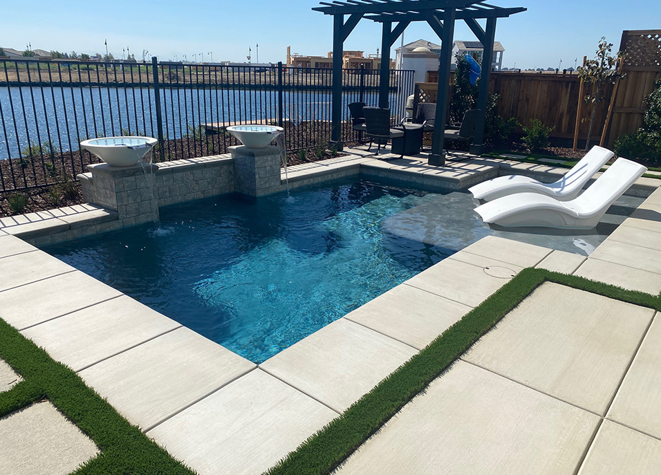 A modern backyard pool with two white lounge chairs on a sun shelf, elegant water features, and a pergola seating area.
