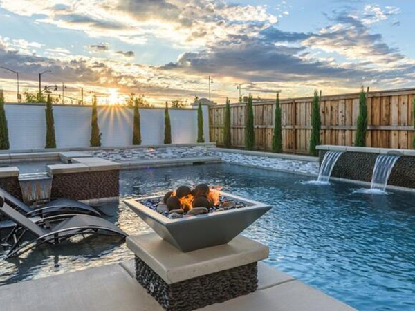 A stylish backyard pool featuring a fire pit, cascading waterfalls, and lounge chairs, surrounded by a wooden fence and lit by the soft glow of the setting sun against a partly cloudy sky.