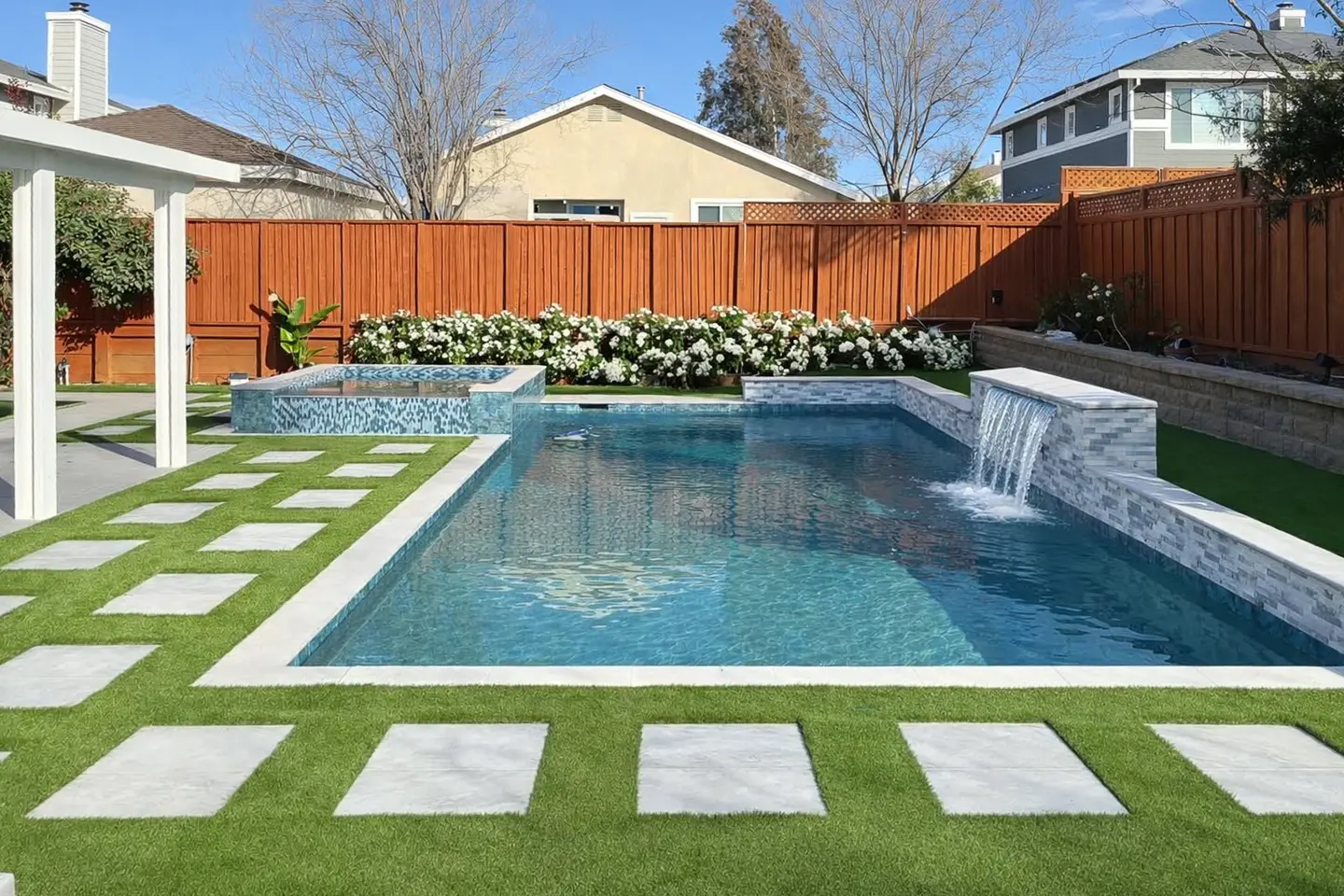Modern backyard swimming pool with an elevated spa, waterfall feature, artificial turf, and concrete stepping stones.